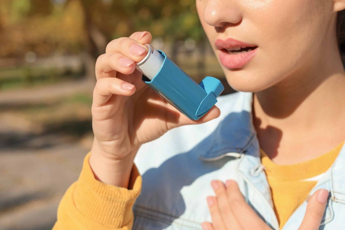 Woman holding a blue reliever inhaler to her mouth with her hand on her chest, showing thunderstorm asthma symptoms outdoors